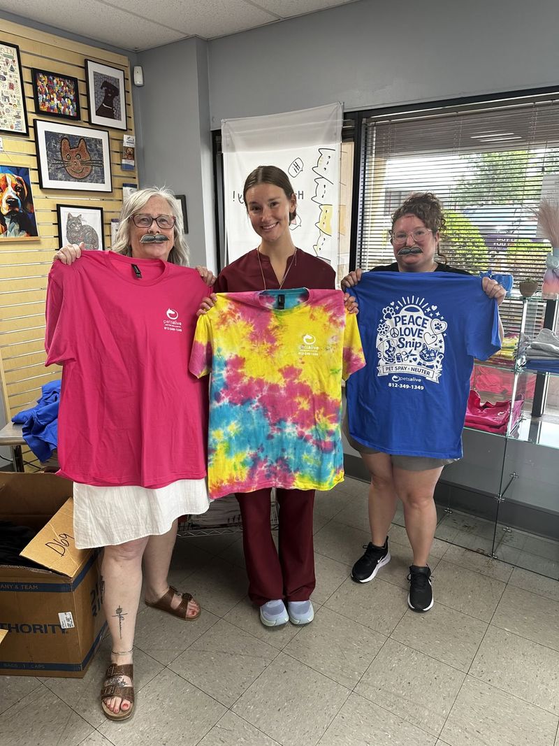 Three smiling Pets Alive team members holding up colorful tie-dye clinic t-shirts